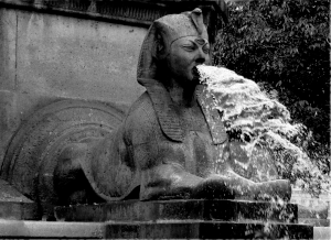 a grayscale photo of the sphinx detail on the fountain of palmier, spewing water from its mouth. it has been edited to have three eyes.