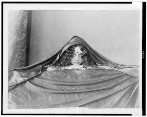 a grayscale photo of a woman lying under a cloth, arms folded under her chin, facing camera. she has curly hair, an archaic smile, and has been edited to have three eyes.